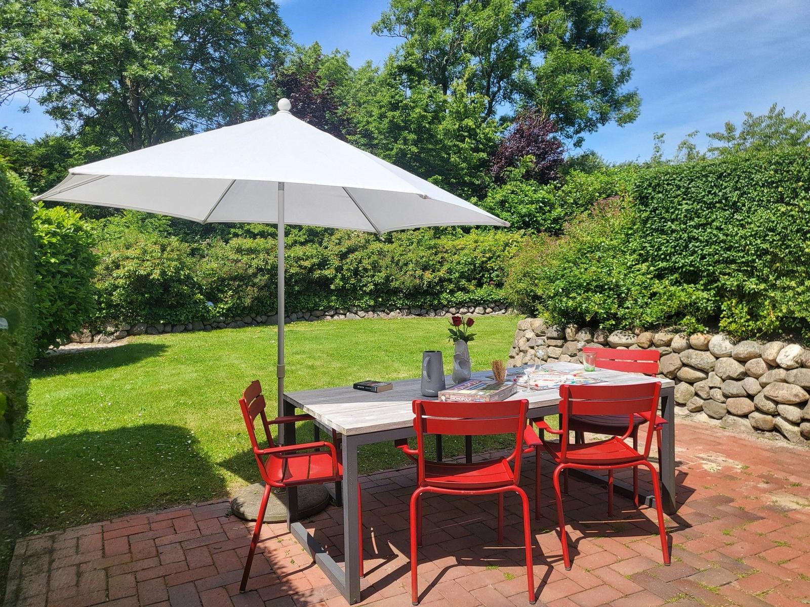 Roter Tisch und Stühle unter weißem Sonnenschirm auf Steinplatten im Garten.