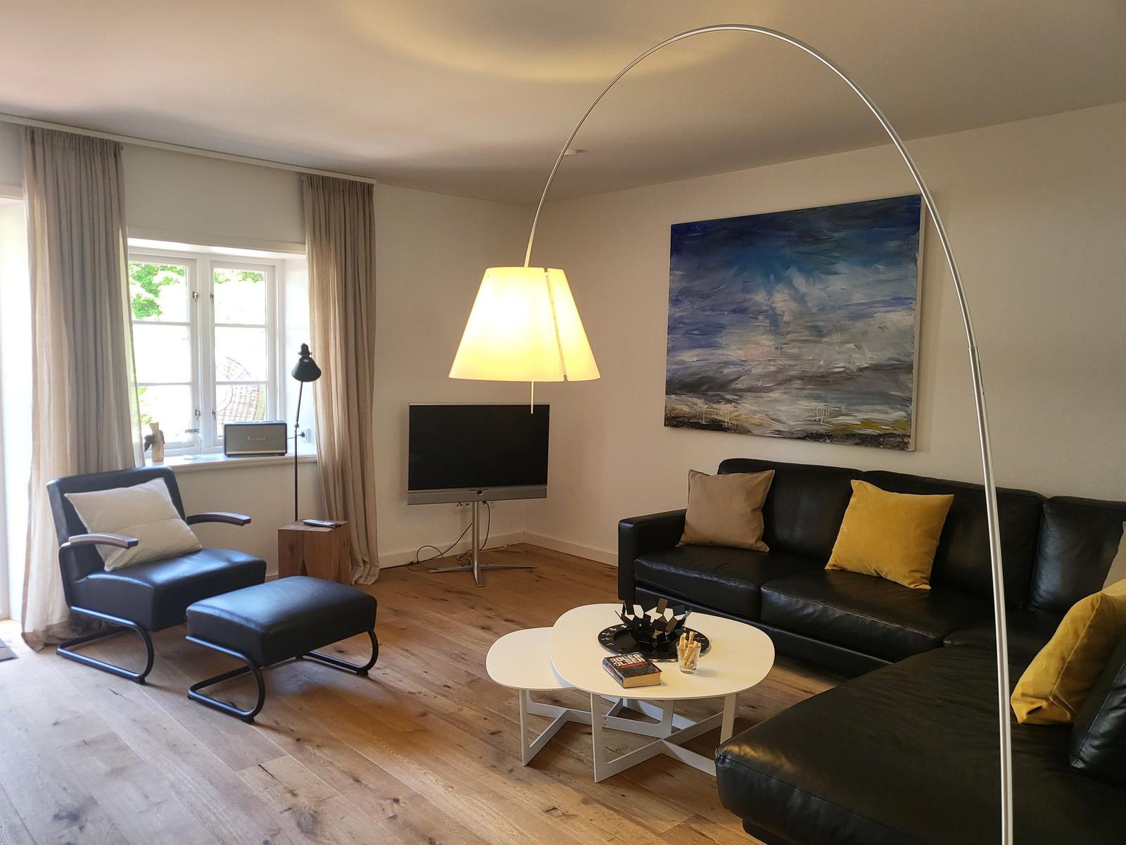 Wohnzimmer mit Sofa, Sessel, Couchtisch und Fernseher. Große Fenster und helle Holzböden.