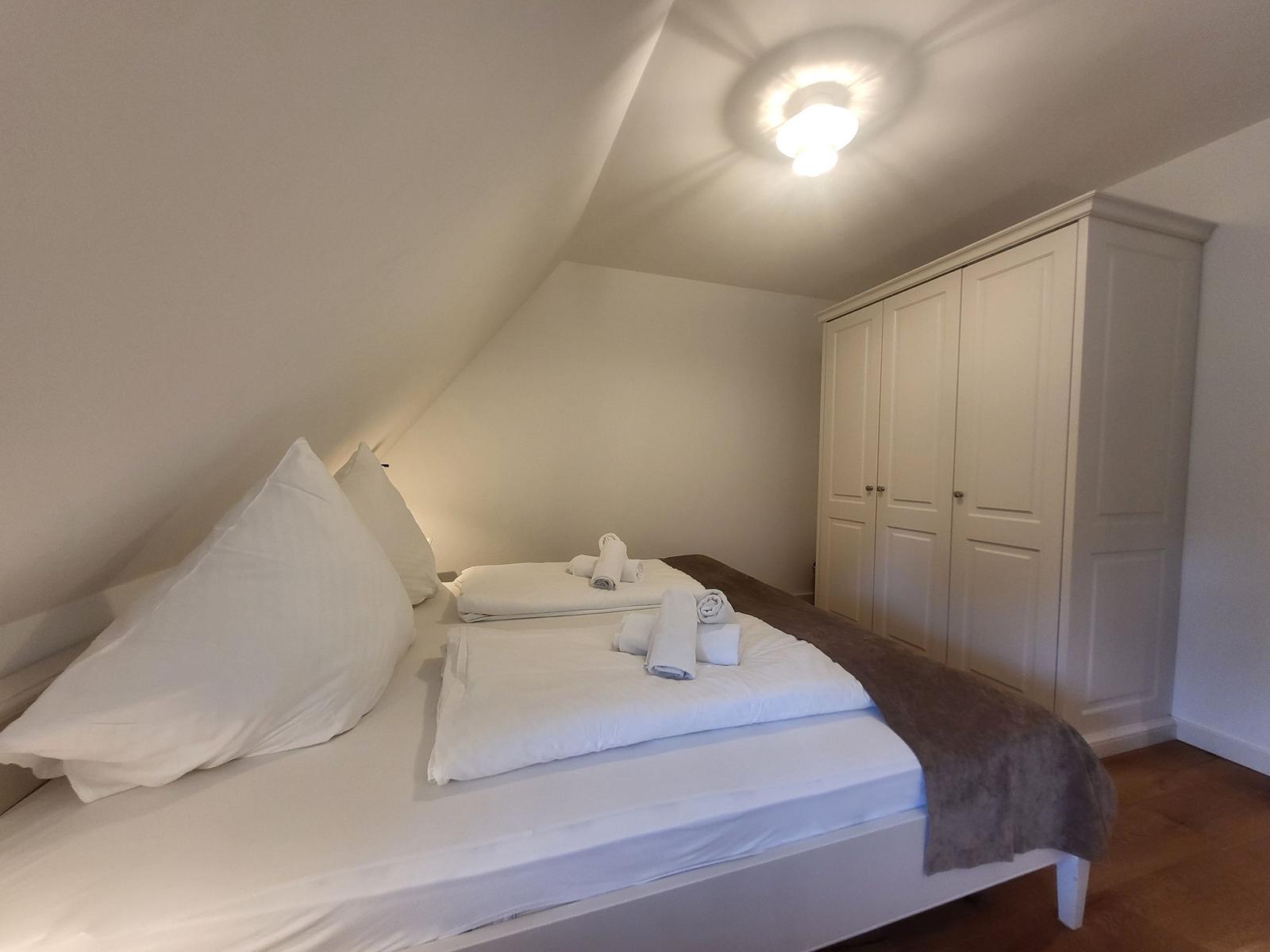 Schlafzimmer mit Dachschrägen, Bett mit weißen Kissen und Handtüchern, weißer Kleiderschrank.