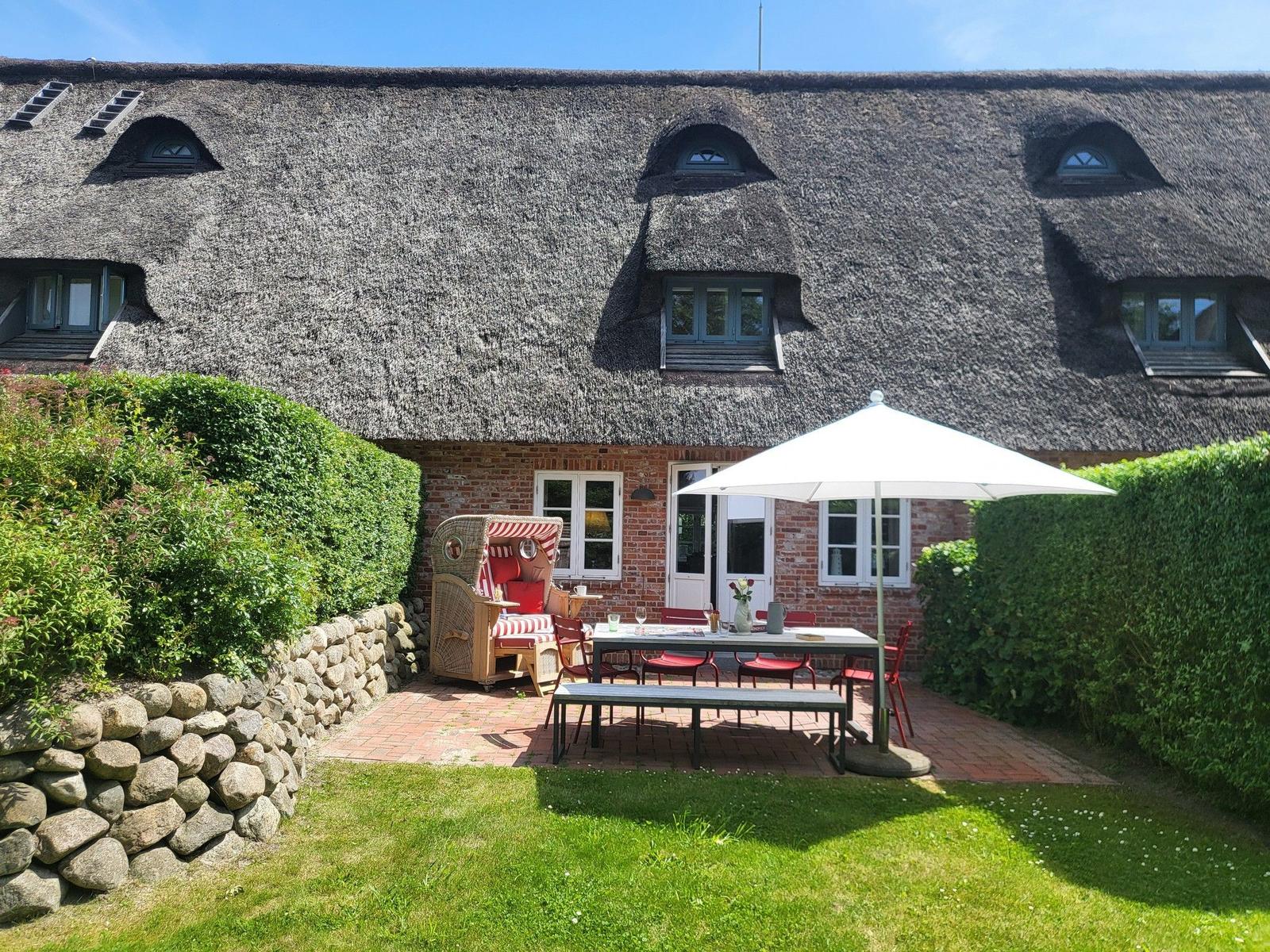 Hinterhof mit Tisch, Stühlen und Sonnenschirm vor einem Strohdachhaus mit Fenstern.