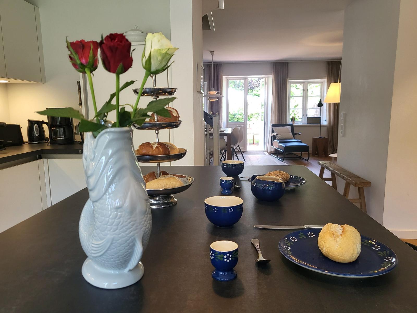 Esstisch mit Blumenvase, Tassen und Brot. Im Hintergrund Küche und Wohnbereich.