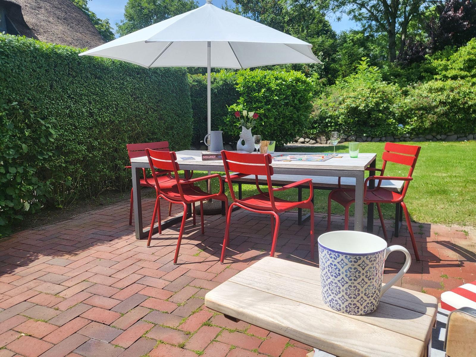 Roter Gartenbereich mit Tisch, Stühlen und Sonnenschirm. Kaffeebecher auf Holztisch.
