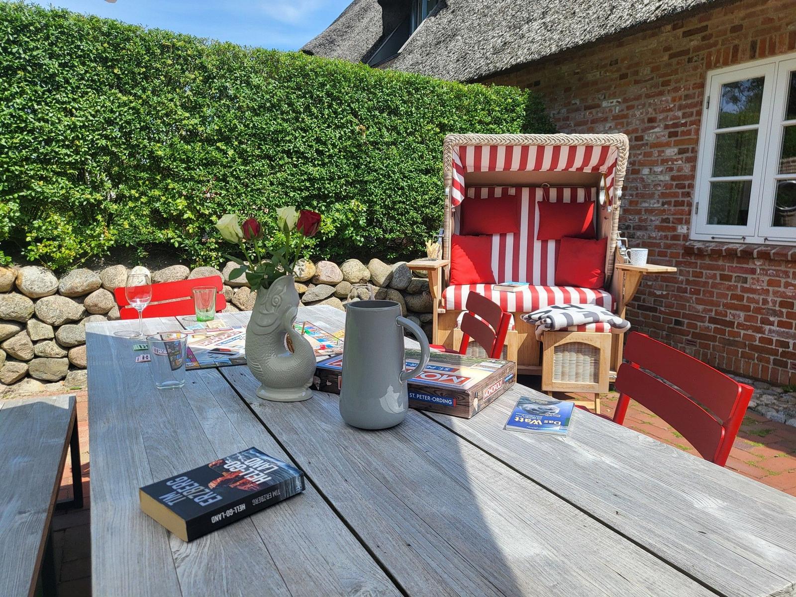 Holztisch mit Buch, Krug und Blumen vor Strandkorb und Backsteinhaus.