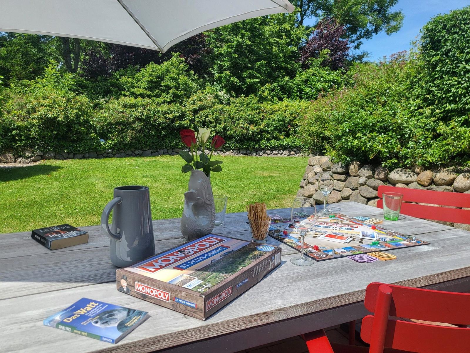 Holztisch mit Monopoly, Wein und Blumen unter Sonnenschutz im Garten.