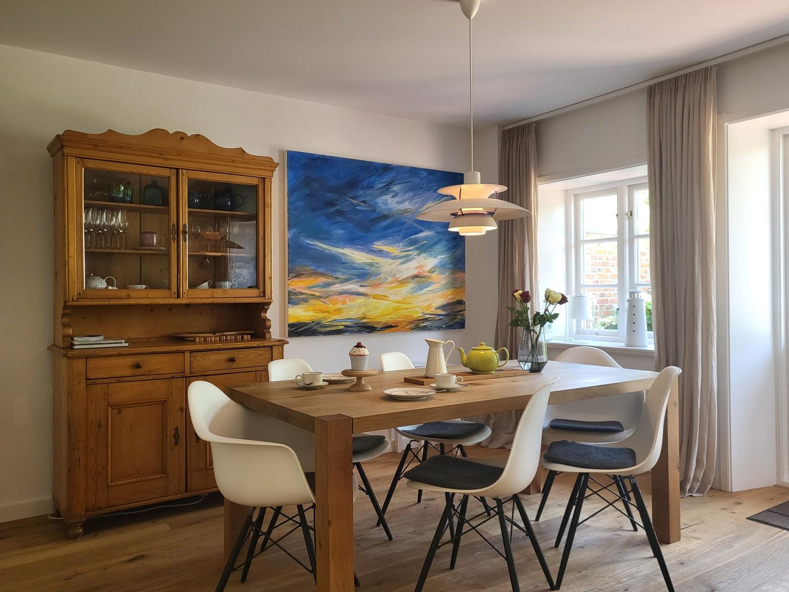 Esszimmer mit Holztisch, weißen Stühlen und Holzschrank. Wandbild und Fenster mit Vorhängen.