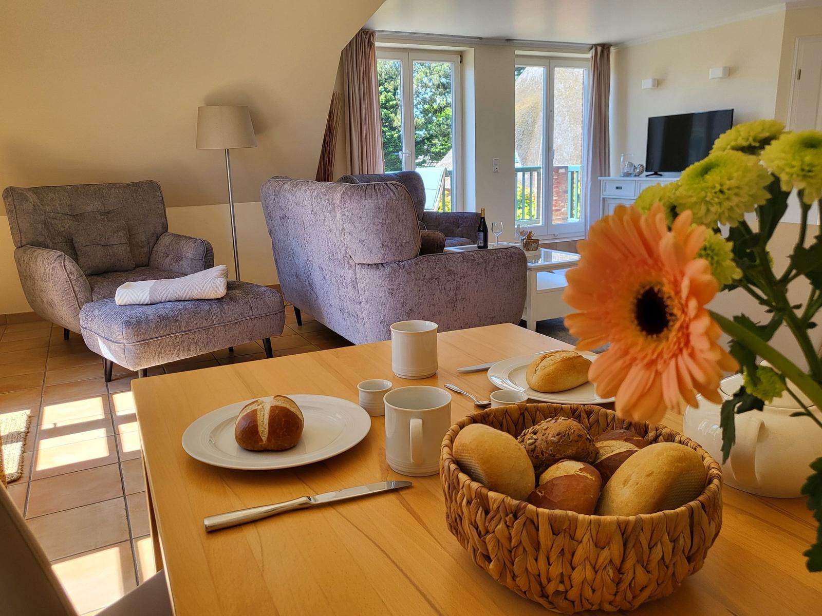 Esszimmer mit Holztisch, Brot und Blumen. Im Hintergrund Wohnzimmer mit Sesseln und Fenstern.