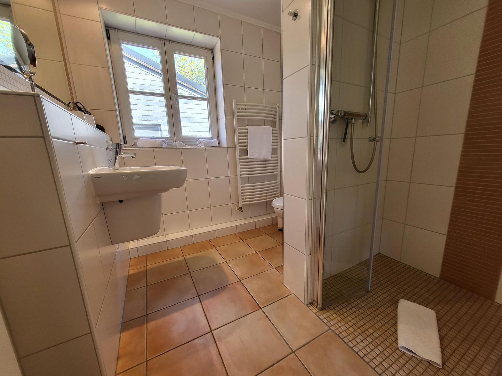 Badezimmer mit Dusche, Waschbecken und Fenster. Fliesenboden und Heizung.