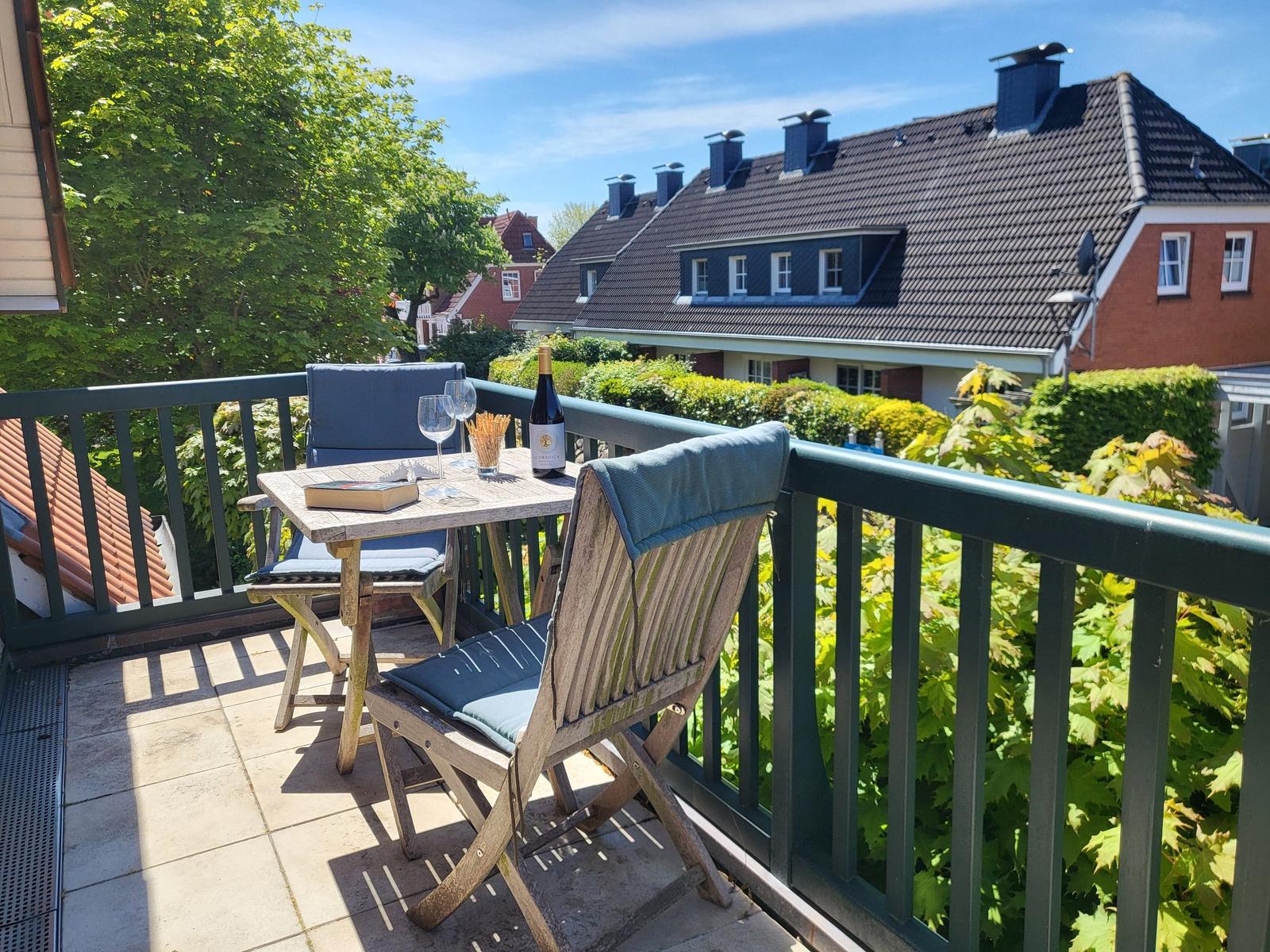 Terrasse mit Tisch, zwei Stühlen, Weinflasche und Buch. Blick auf Wohnhäuser und grüne Bäume.