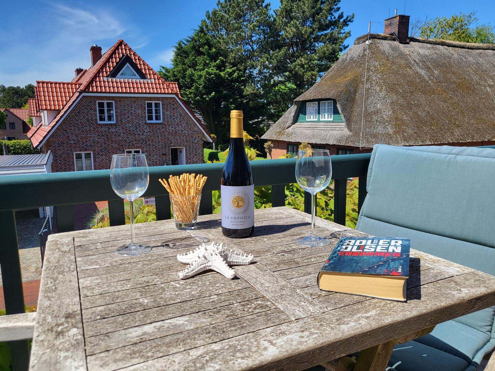 Holztisch mit Wein, Gläsern, Kräckern und Buch auf Terrasse mit Blick auf Häuser.