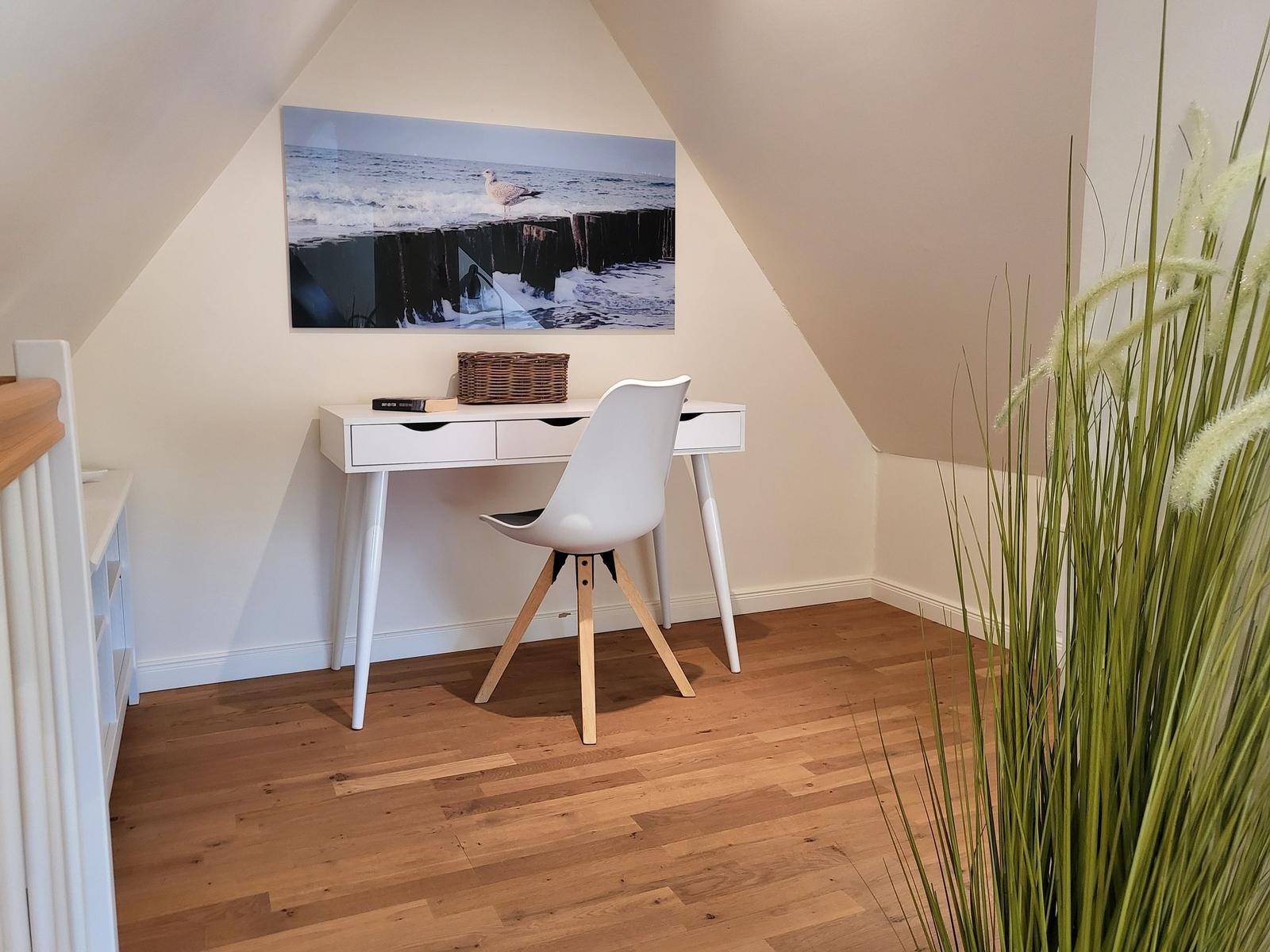 Schreibtisch mit Stuhl unter Dachschrägen, Wandbild und Pflanze im Vordergrund.