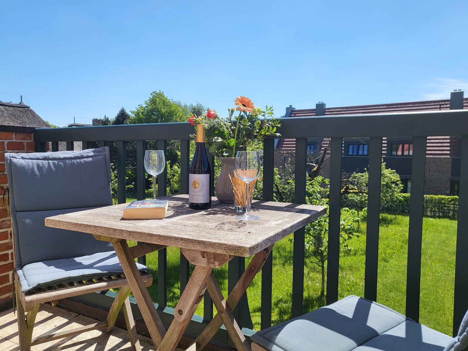Terrasse mit Tisch, Stühlen, Wein und Blumen. Blick auf grünen Garten und Häuser.