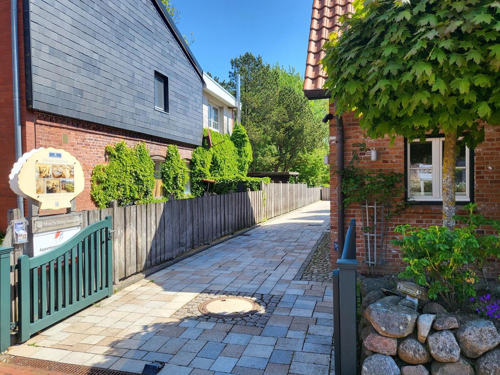 Pflasterweg führt durch einen Garten mit Steinmauer und Bäumen zu einem Haus.