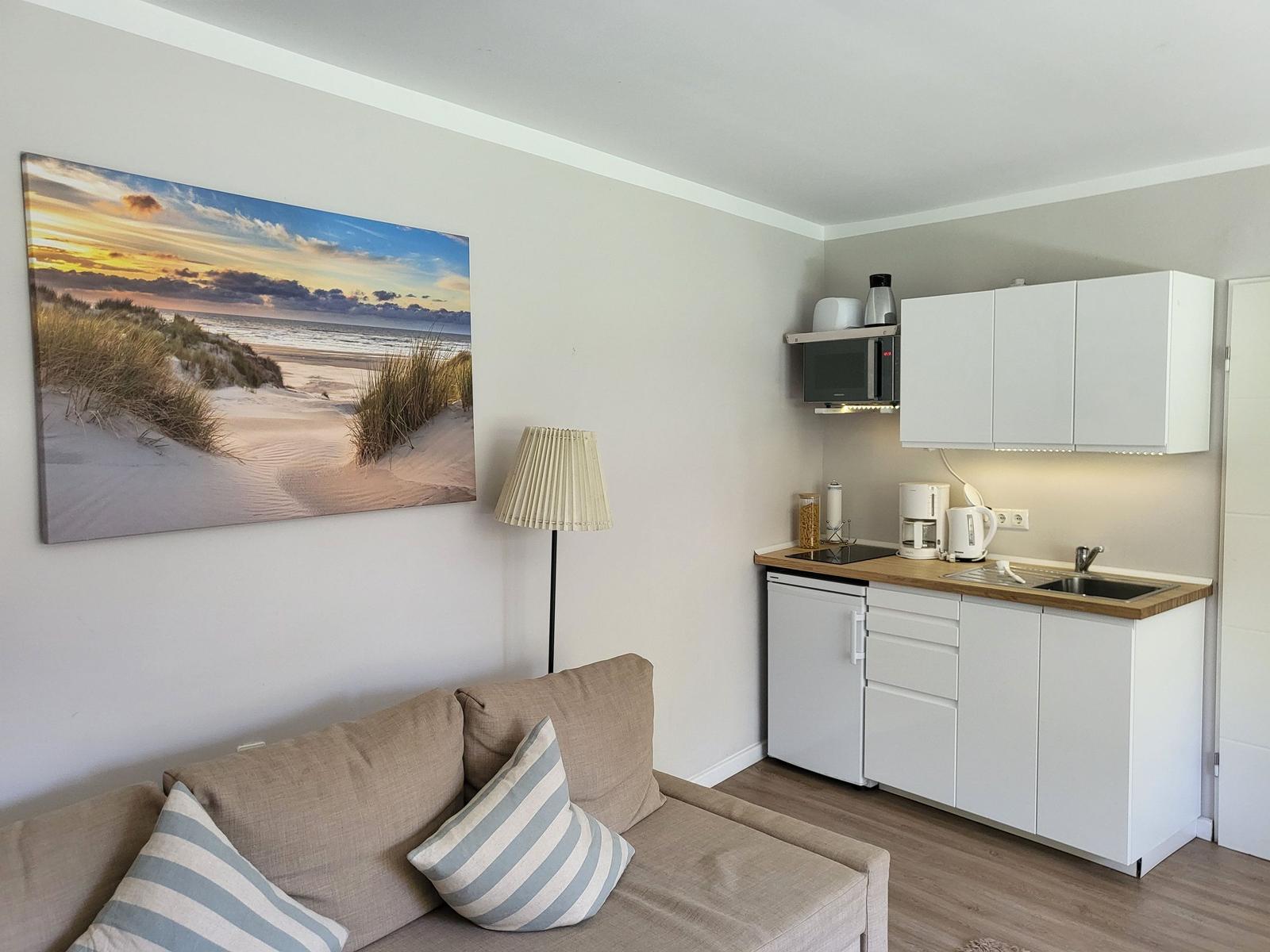 Wohnbereich mit Sofa, Küchenzeile und Wandbild von Strand.