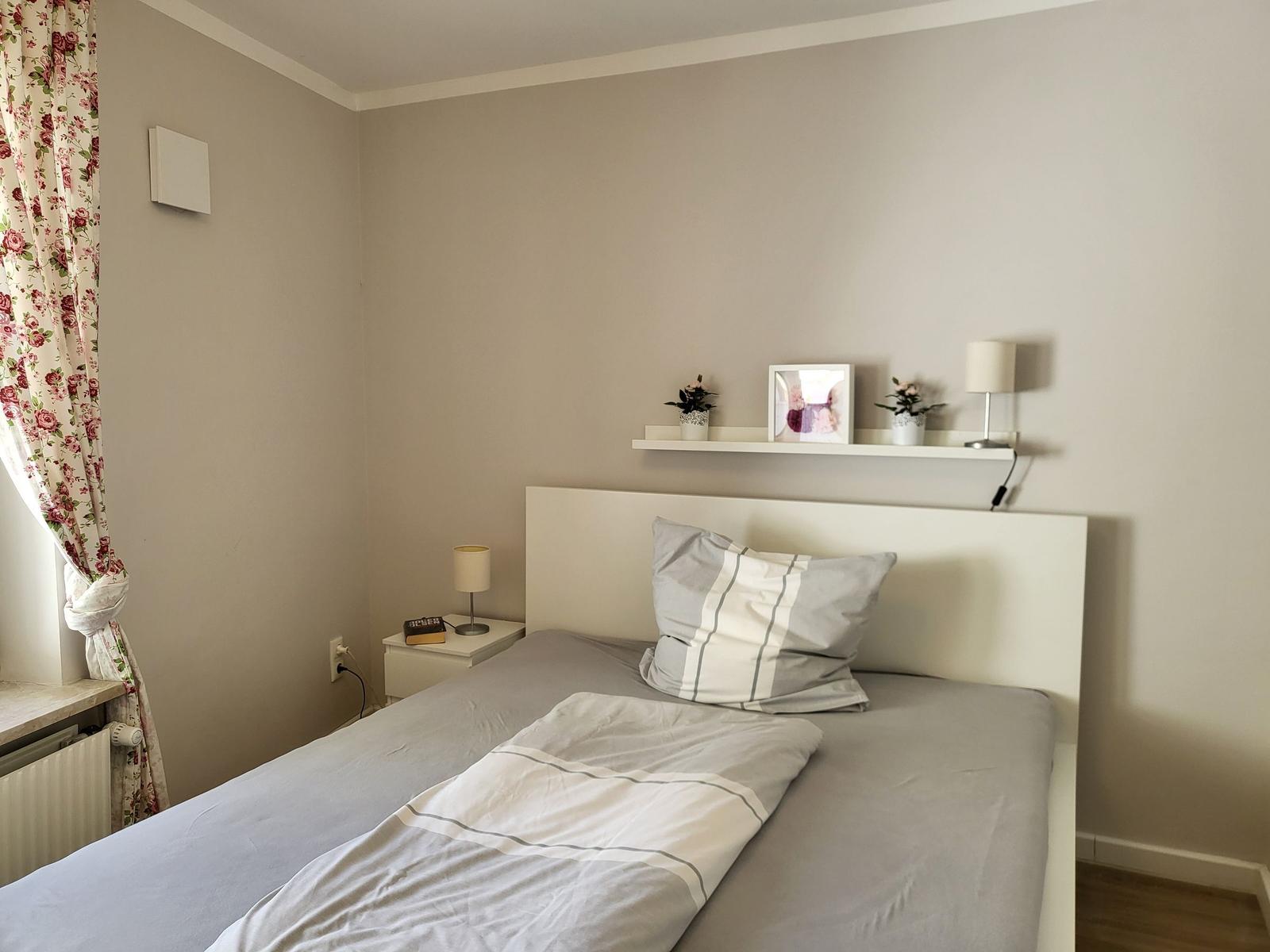 Schlafzimmer mit Bett, Nachttisch und Wandregal mit Pflanzen und Foto.