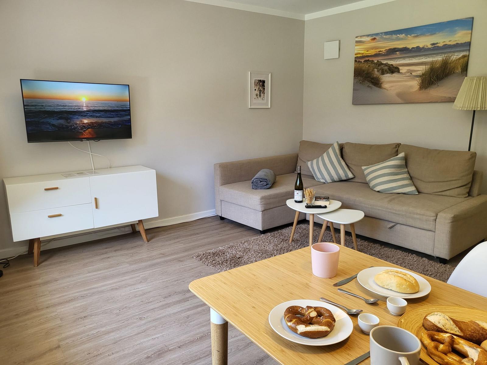 Wohnzimmer mit Sofa, TV und Esstisch mit Brötchen.