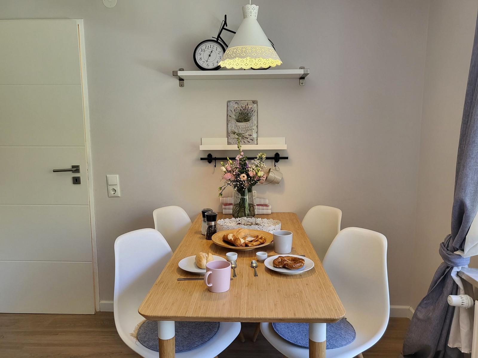 Esstisch mit vier Stühlen, Blumen und Kaffeetassen. Wandregale mit Uhr und Lampe.