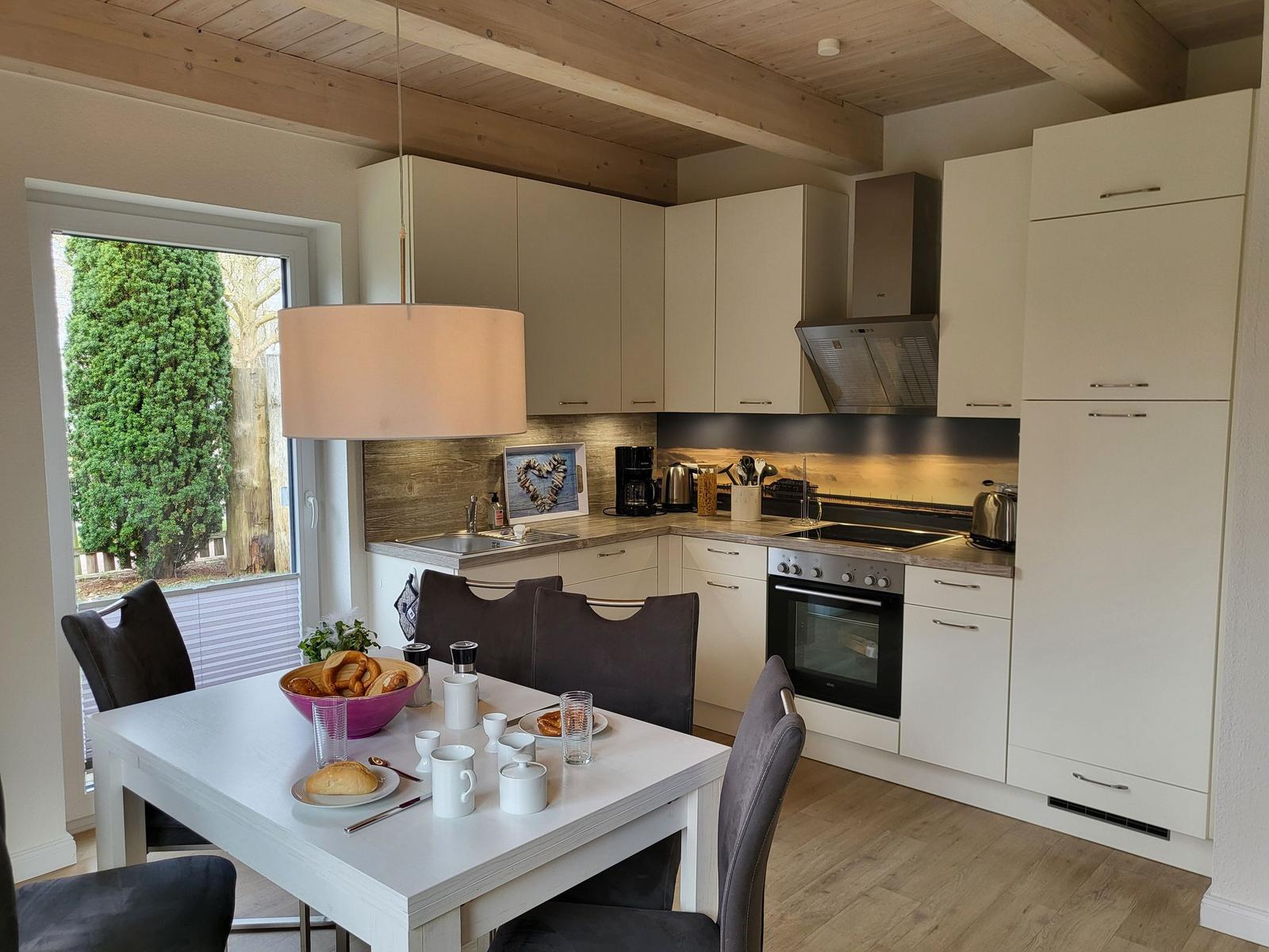 Küche mit Essbereich, weißen Schränken und Holzdecke. Tisch mit Frühstück und Fenster zum Garten.