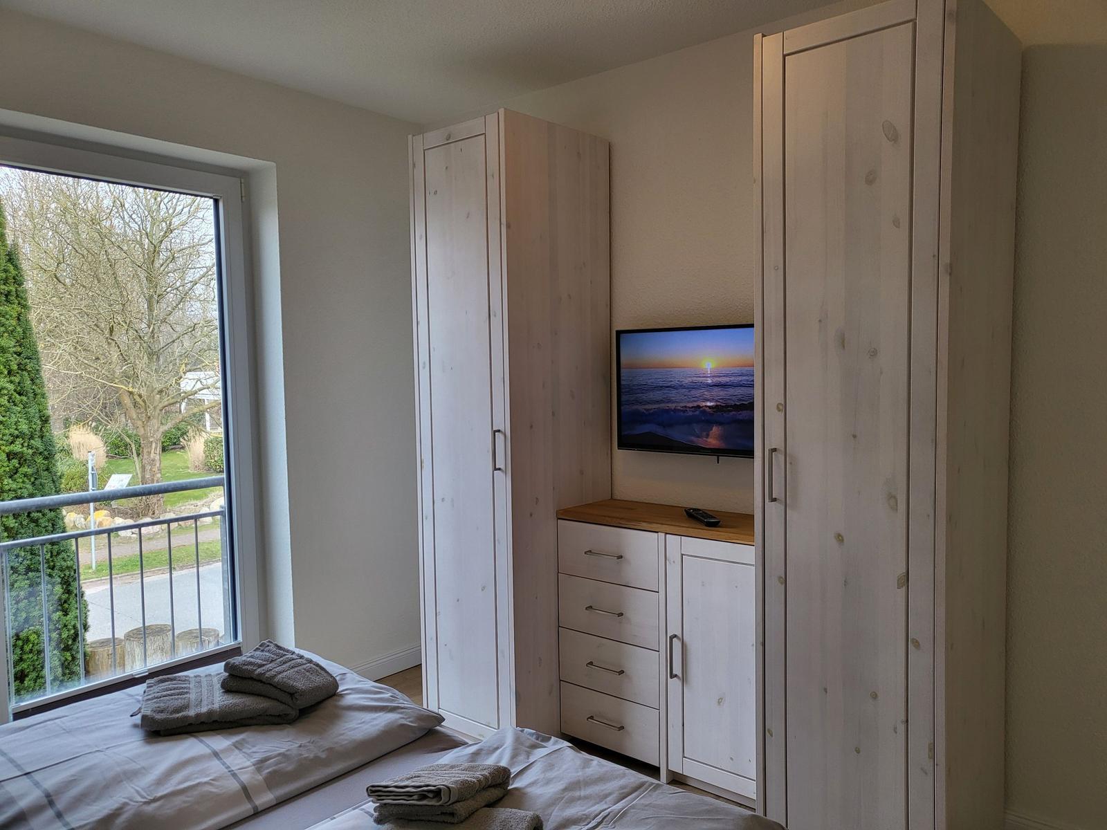 Schlafzimmer mit Bett, Kleiderschränken und Fernseher. Balkon mit Blick auf Grünfläche.