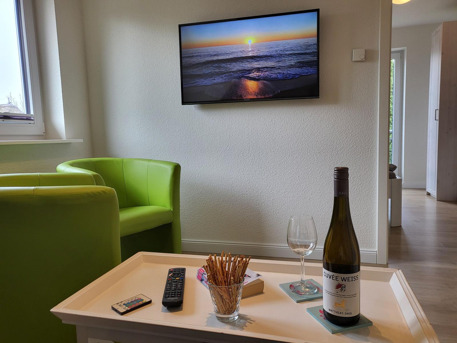 Ein Wohnzimmer mit grünen Sesseln, Fernseher und Tisch mit Wein und Knabberzeug.