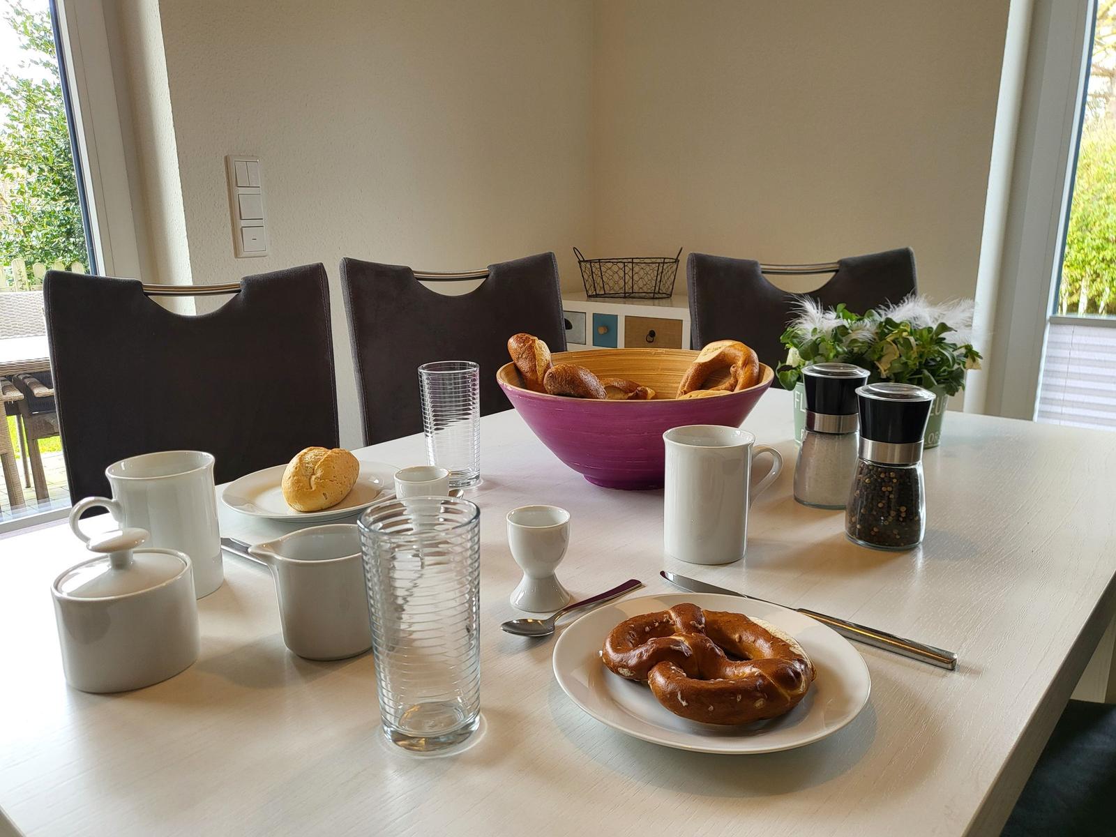Frühstückstisch mit Brot, Kaffee und Gewürzgläsern vor Fenstern.