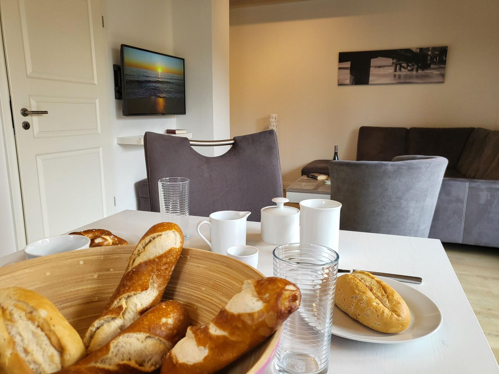 Frühstückstisch mit Brot, Gläsern und Tassen. Im Hintergrund Wohnbereich mit Sofa und Fernseher.