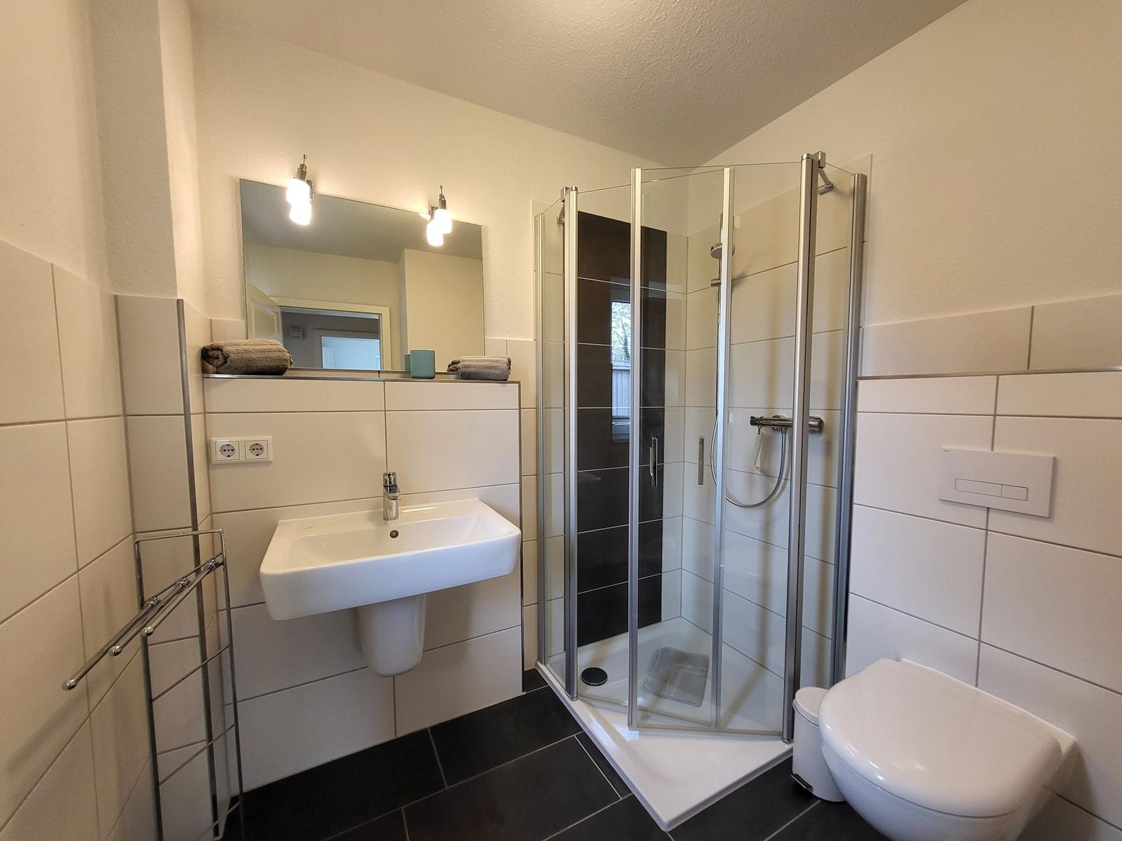 Badezimmer mit Dusche, Waschbecken und Toilette. Fliesenwand und dunkler Boden.