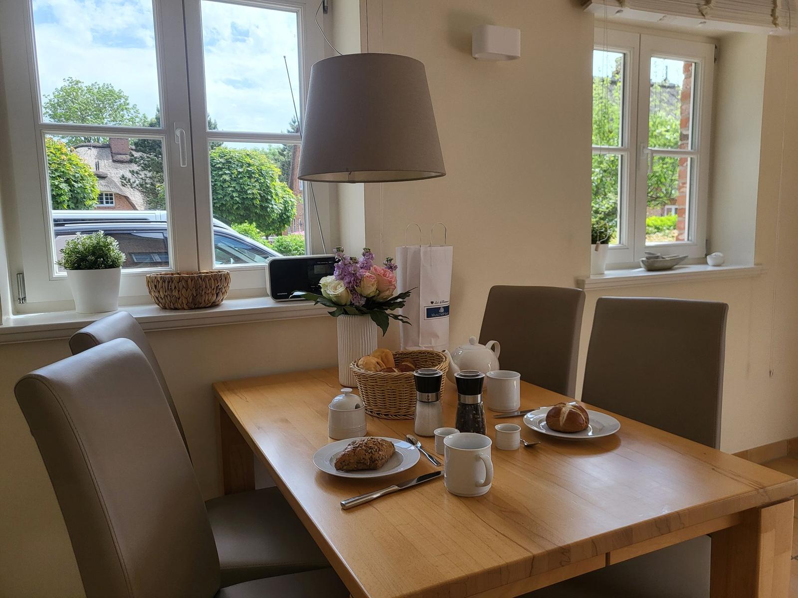 Esszimmer mit Tisch, Stühlen und Fenstern. Auf dem Tisch stehen Tassen und Brötchen.