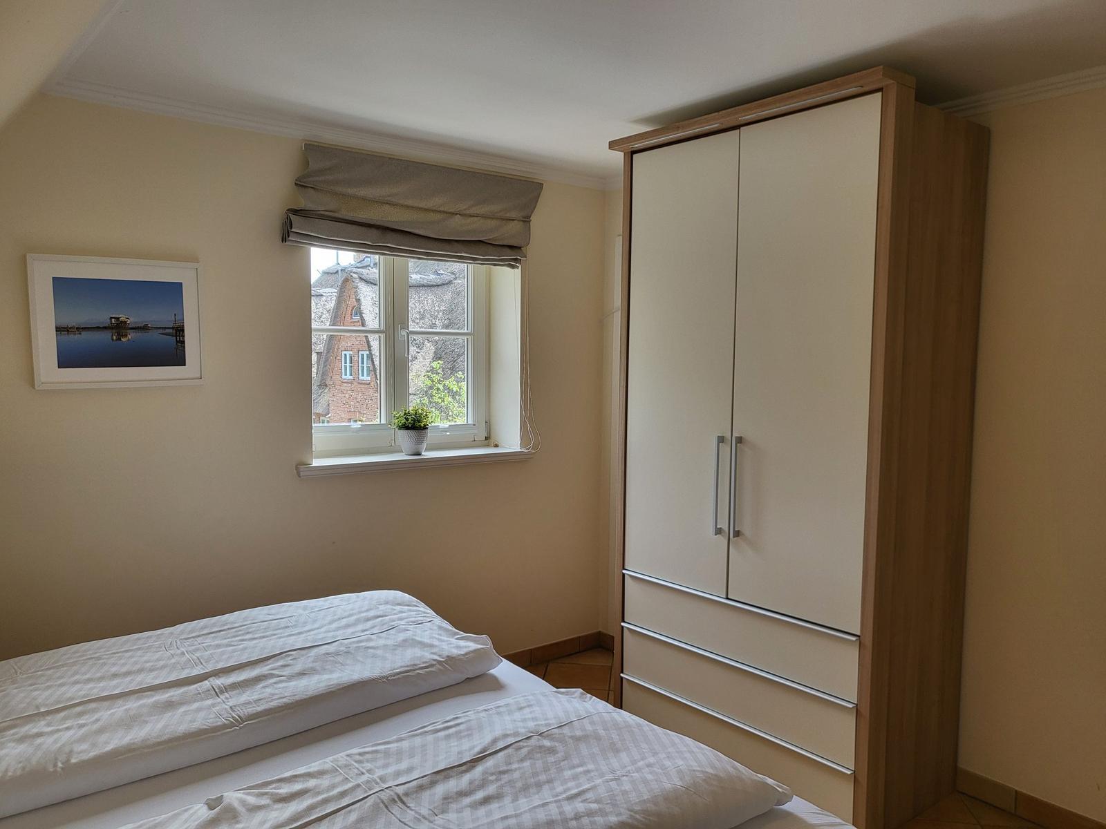 Schlafzimmer mit Bett, Kleiderschrank und Fenster mit Blick auf Dachhäuser.