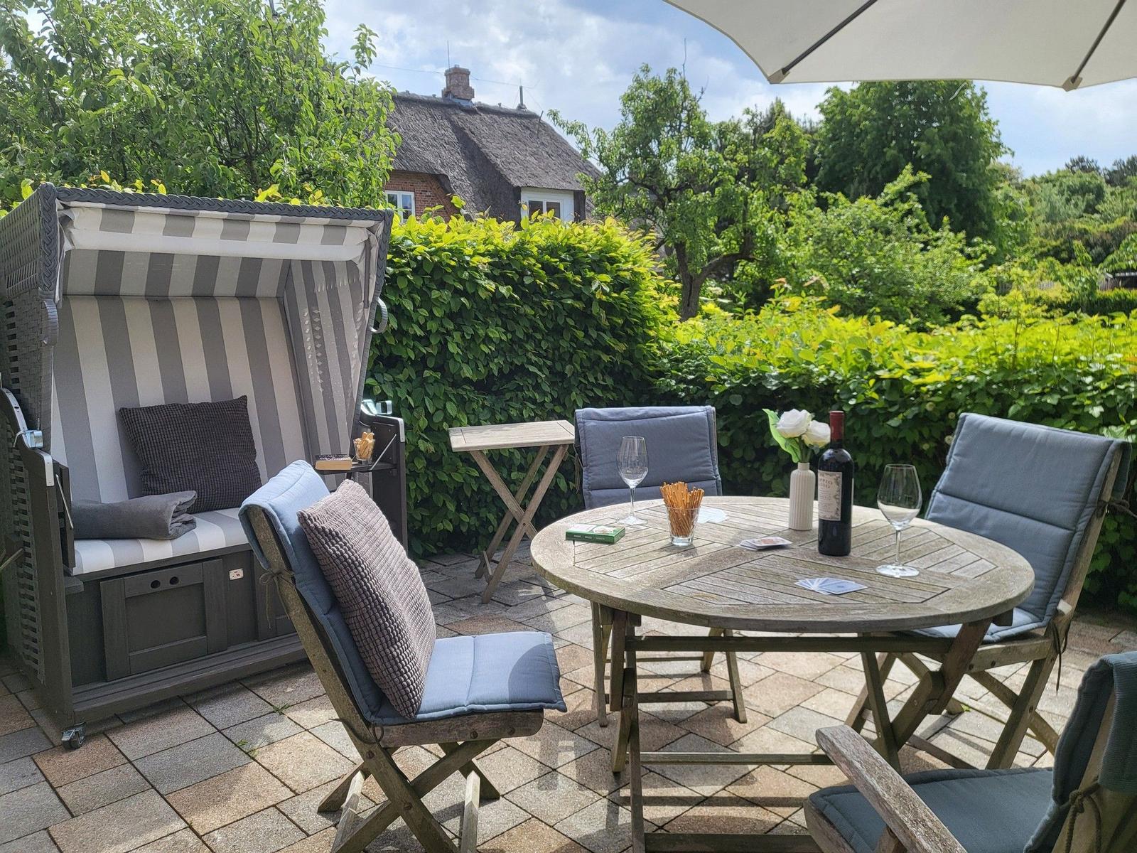 Terrasse mit Tisch, Stühlen und Strandkorb. Hinterhof mit Hecke und Haus im Hintergrund.