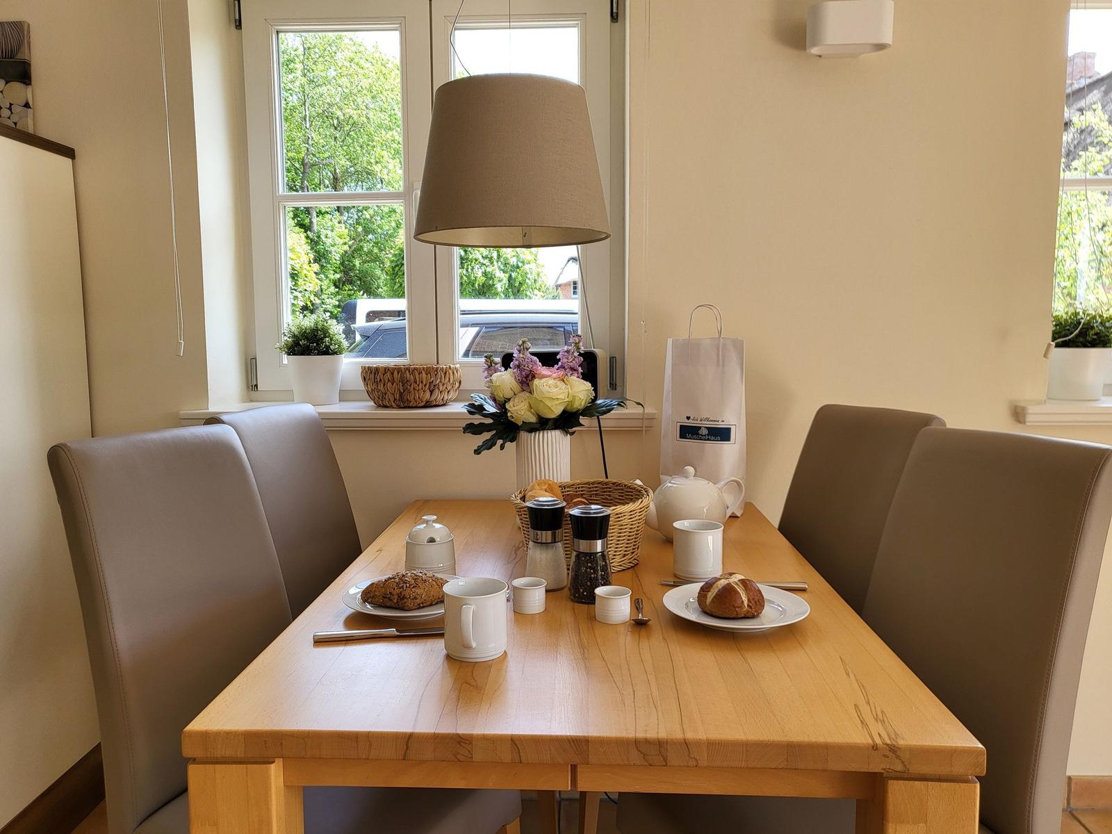 Esstisch mit vier Stühlen, Blumen und Kaffeeutensilien vor Fenster.