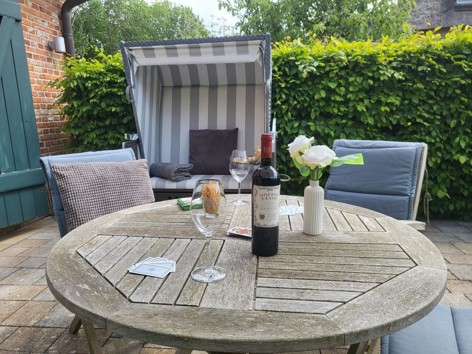 Runder Holztisch mit Wein, Blumen und Spielkarten im Garten mit Liege und Sitzgelegenheiten.