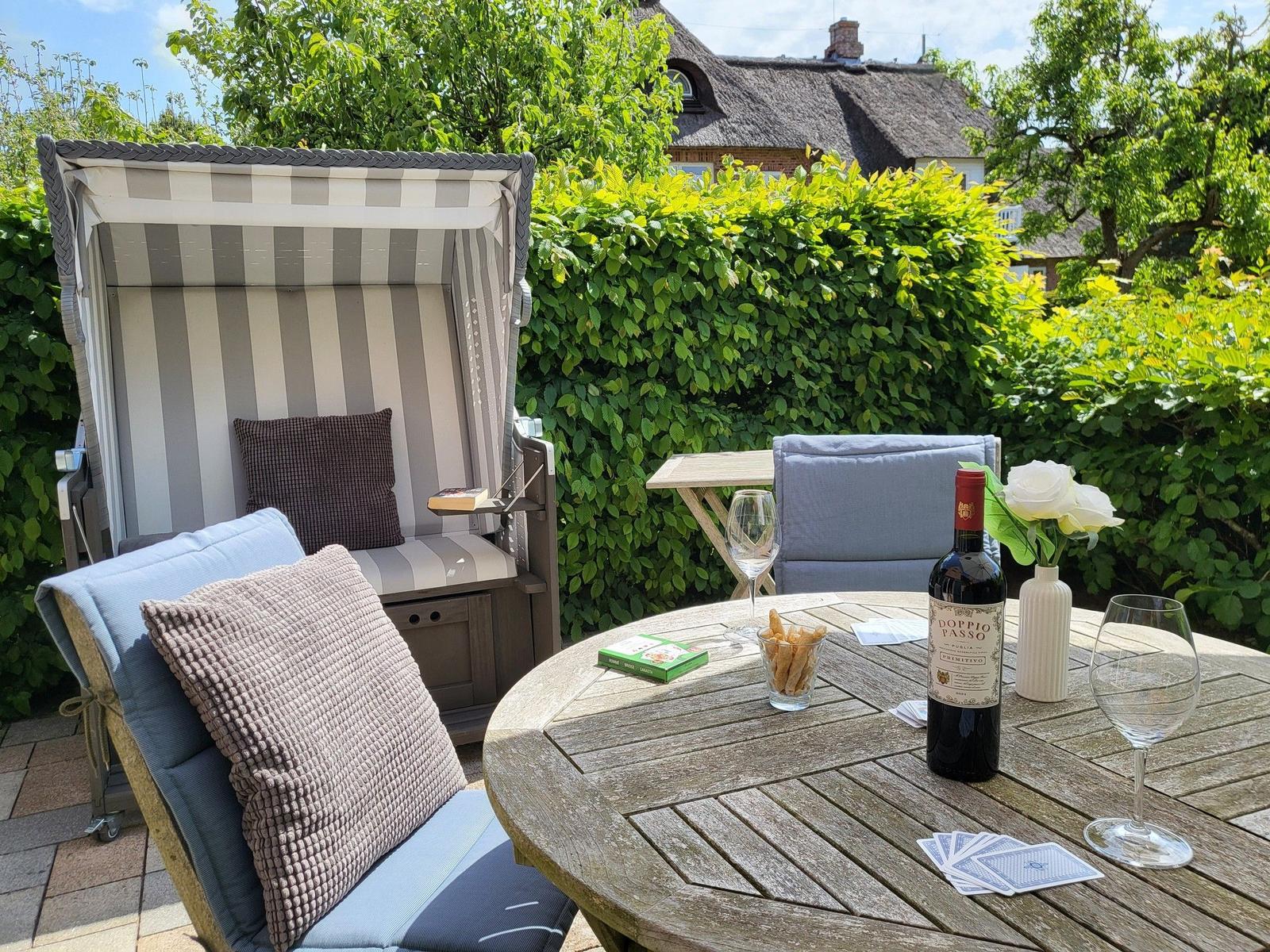 Terrasse mit Strandkorb, Tisch und Stühlen. Weißrosen, Wein und Karten darauf.