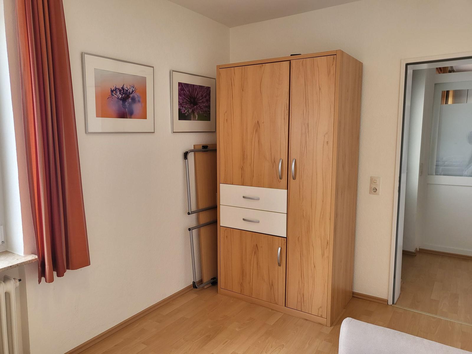 Schlafzimmer mit Holzschrank, zwei Bildern und Vorhang an Fenster.