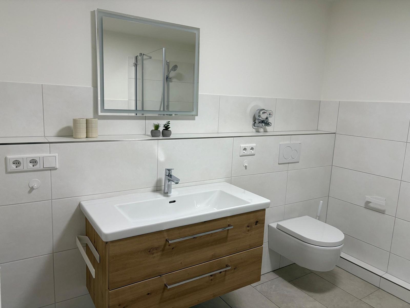 Badezimmer mit Waschbecken, Spiegel und Toilette. Holzschrank und Dusche sichtbar.