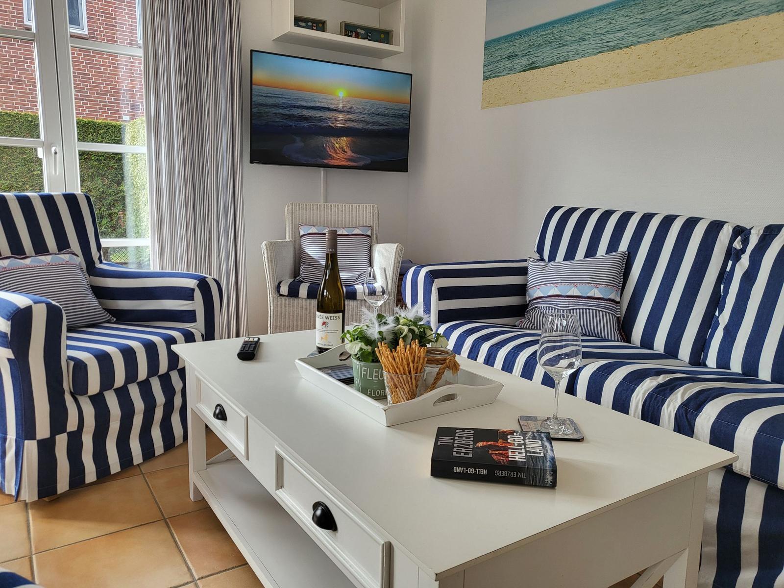 Wohnzimmer mit blau-weiß gestreiften Sitzmöbeln, Fernseher und Couchtisch mit Getränken.