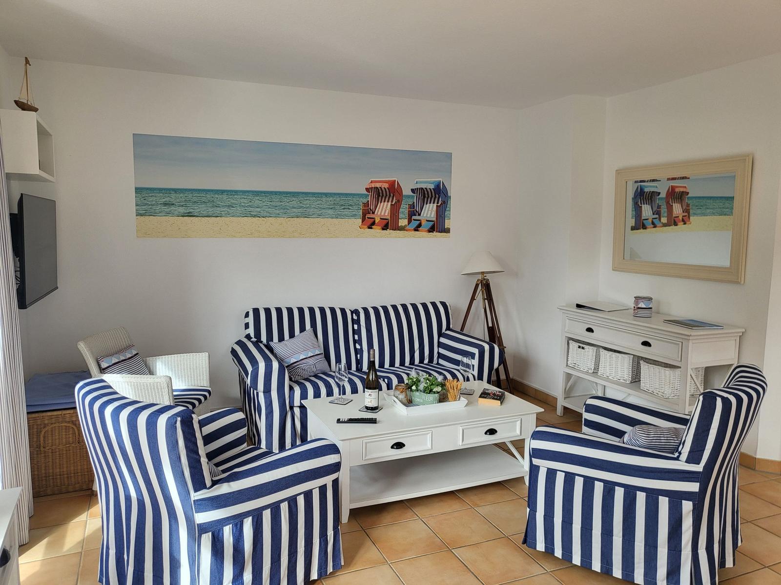 Wohnzimmer mit gestreiften Sitzmöbeln, Couchtisch und Strandmotiven an den Wänden.
