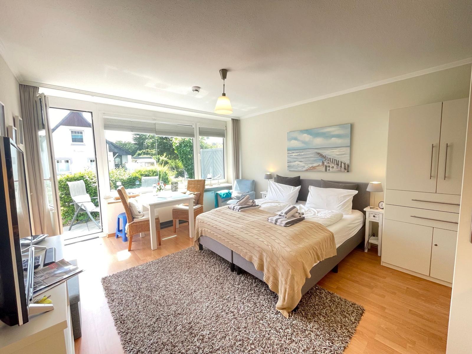 Schlafzimmer mit Bett, Kleiderschrank und Sitzbereich vor großem Fenster.