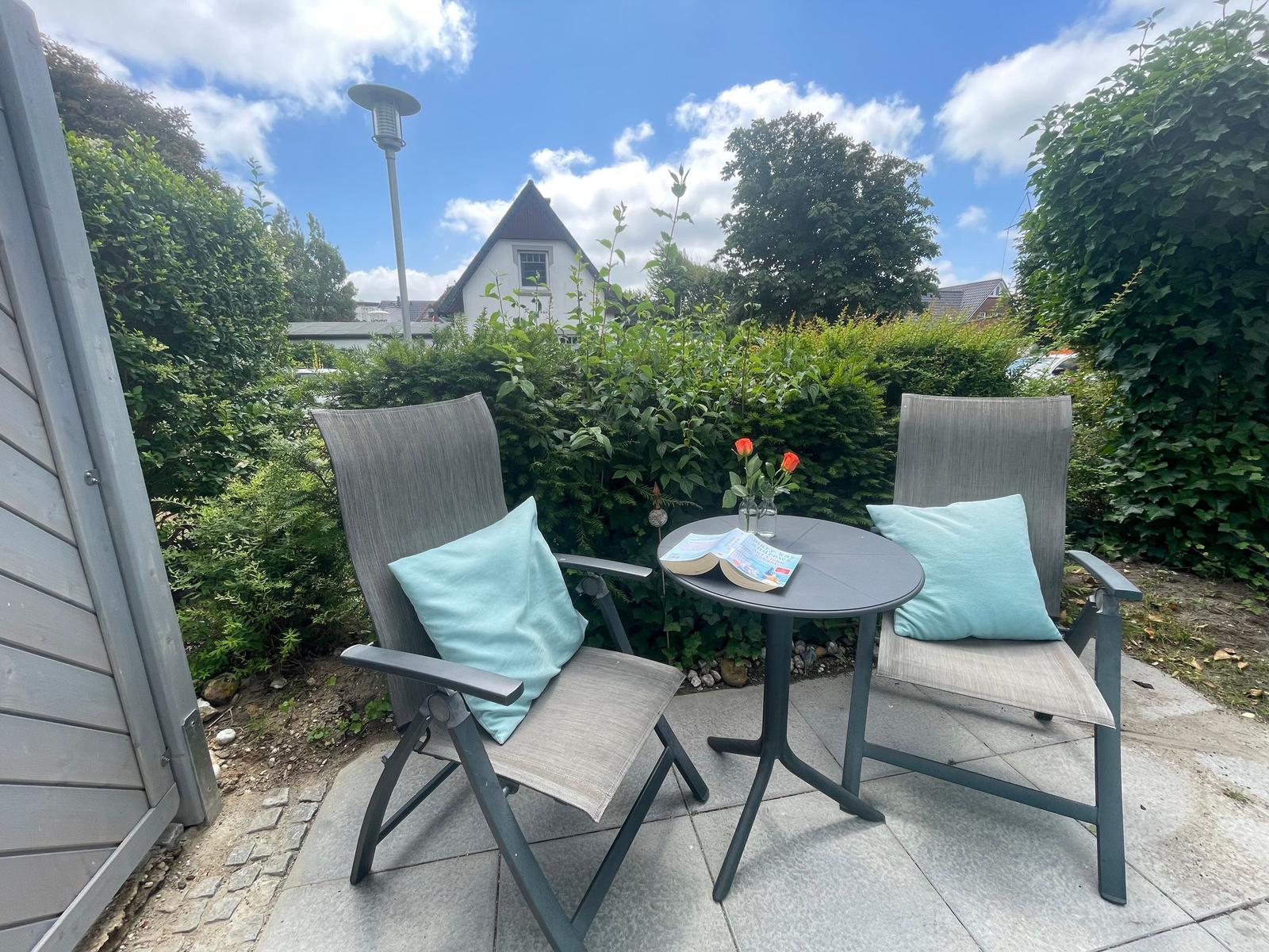 Terrasse mit zwei Stühlen, Tisch und Kissen. Hintergrund: grüne Hecke und Haus.