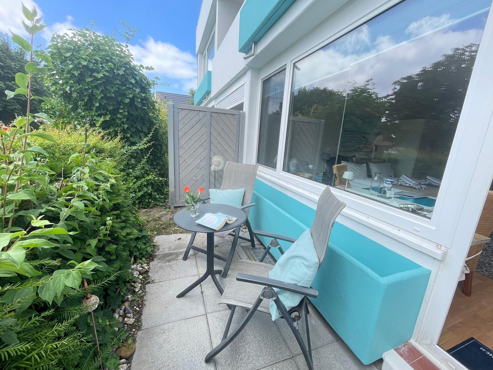 Terrasse mit Tisch, zwei Stühlen und Blick ins Innere. Umgeben von grüner Vegetation.