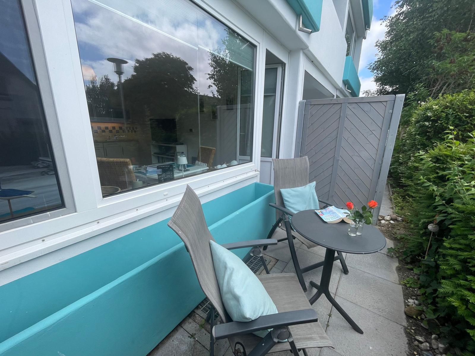 Terrasse mit zwei Stühlen, Tisch und Blumen. Große Fensterfront und grüne Hecke.