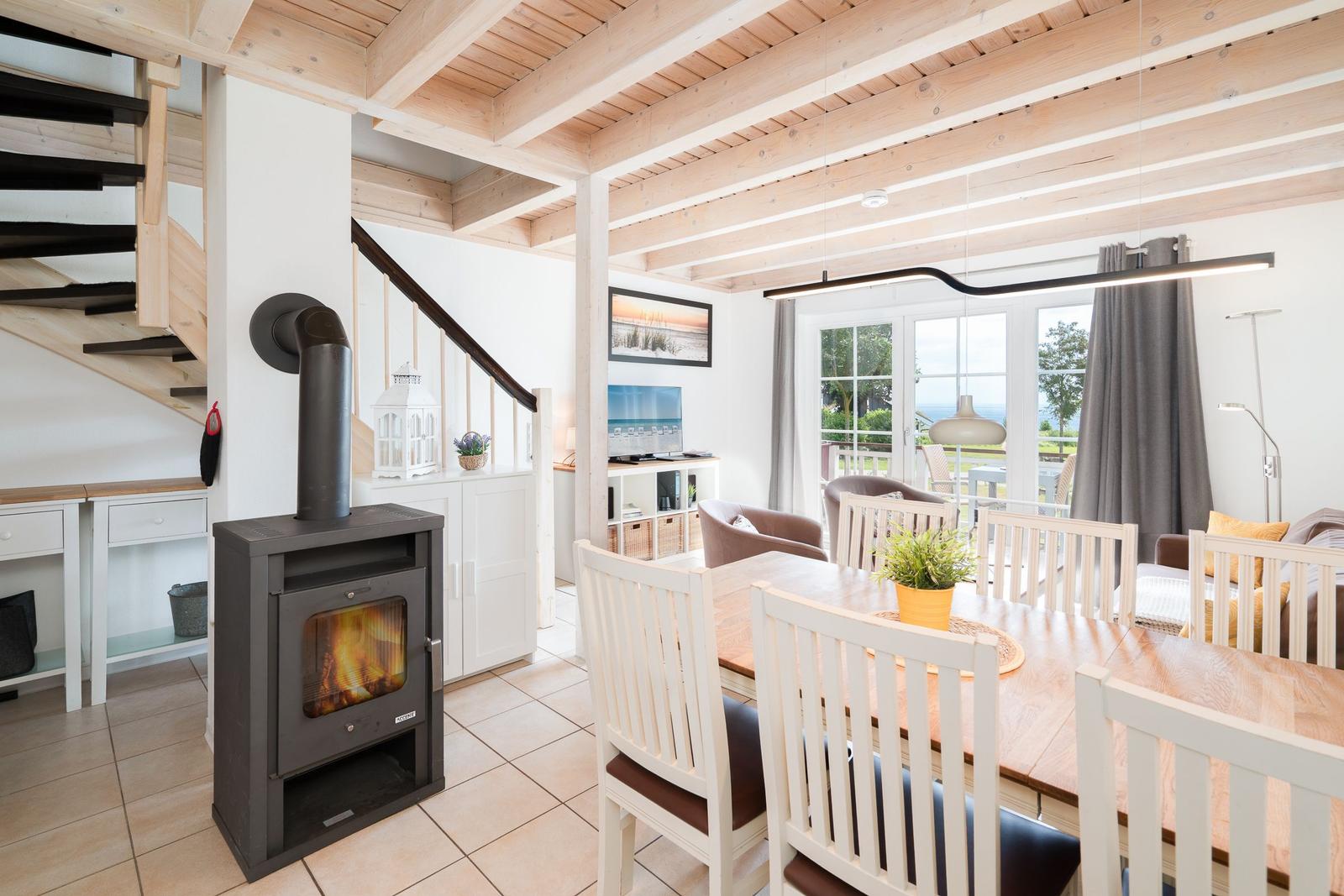 Dining room with wooden table, chairs, and wood stove. Passage to living kitchen with terrace view.