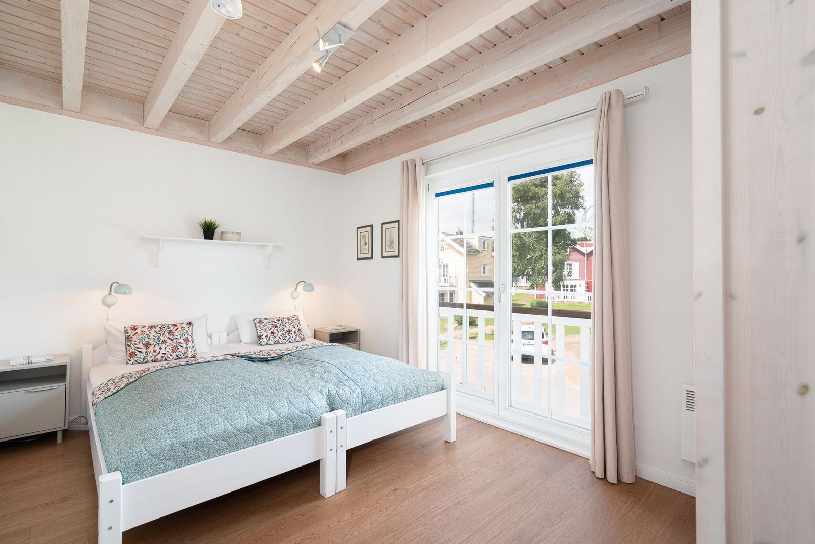 Bedroom with white bed, wooden floor, and balcony door. Ceiling with wooden beams.