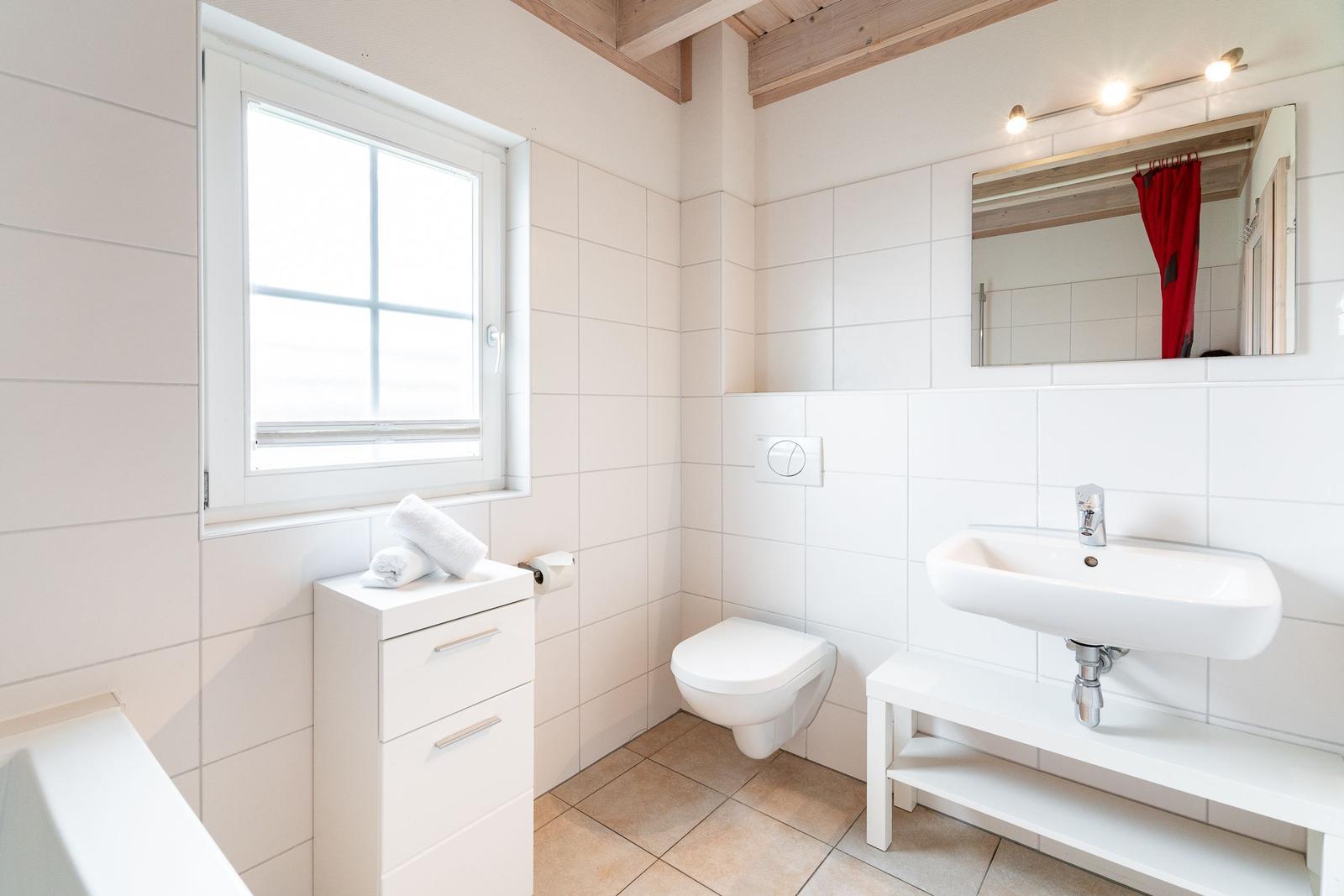 Bathroom with sink, toilet, and window. White tiles and wooden beams.