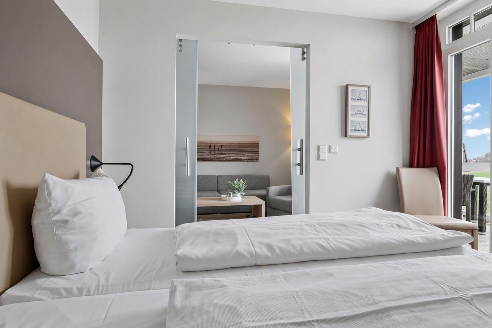 Schlafzimmer mit Bett, Sicht auf Wohnbereich und Balkon mit roten Vorhängen.