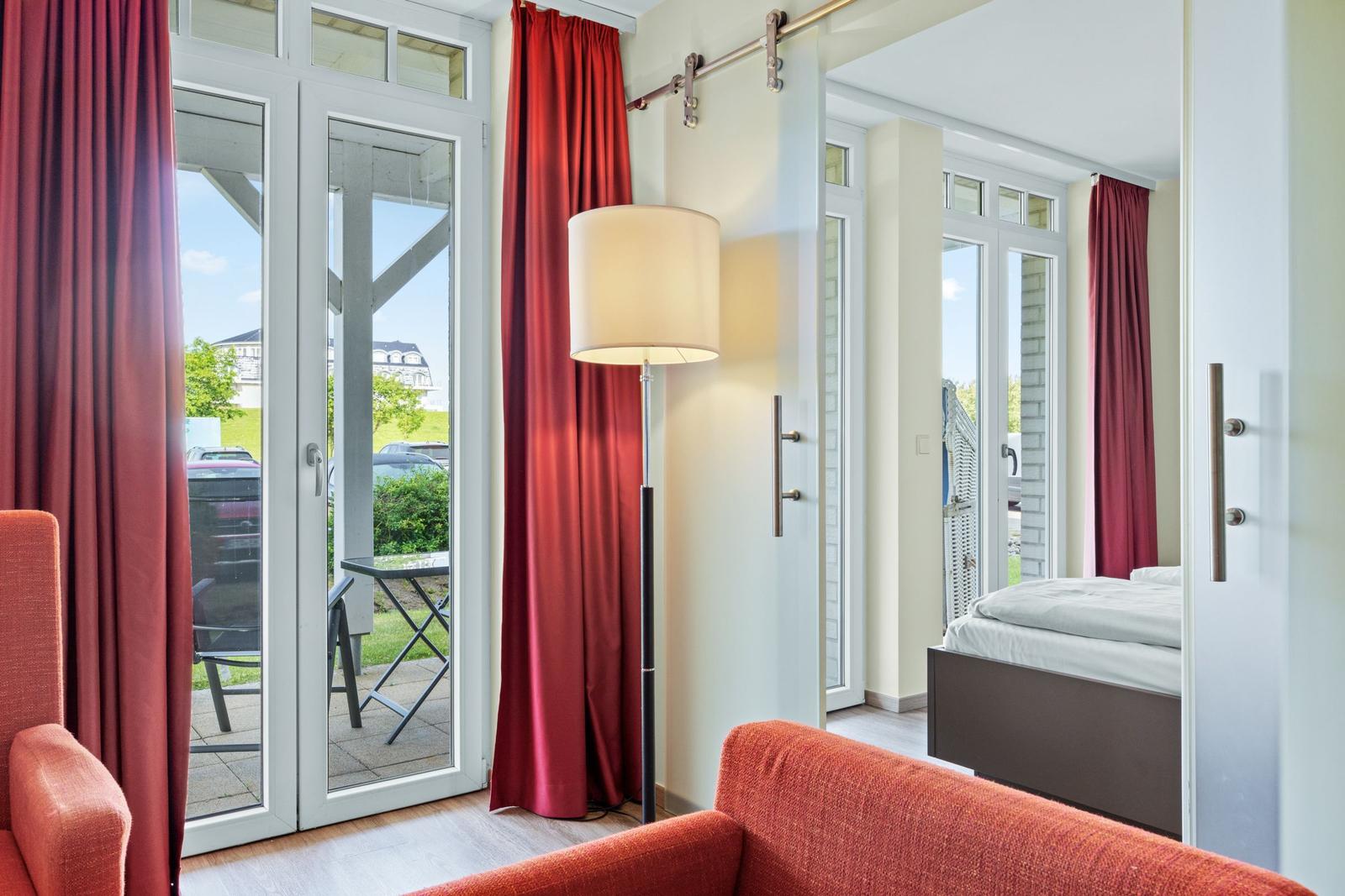 Zimmer mit Schlafbereich und Terrasse. Rote Vorhänge, Stehlampe und Sessel.