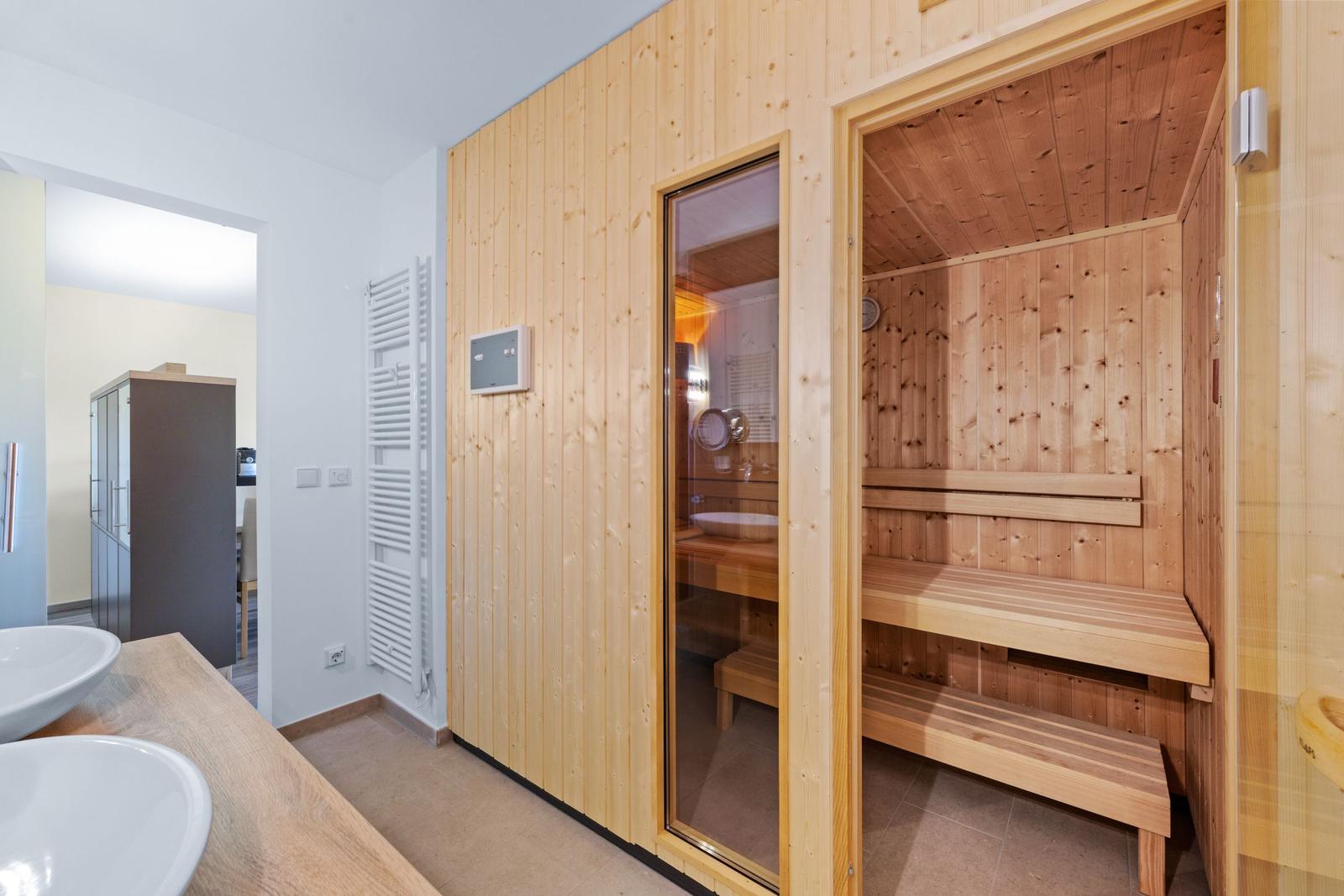 Sauna mit Holzbank und Glaswand, daneben Badewanne und Waschbecken.