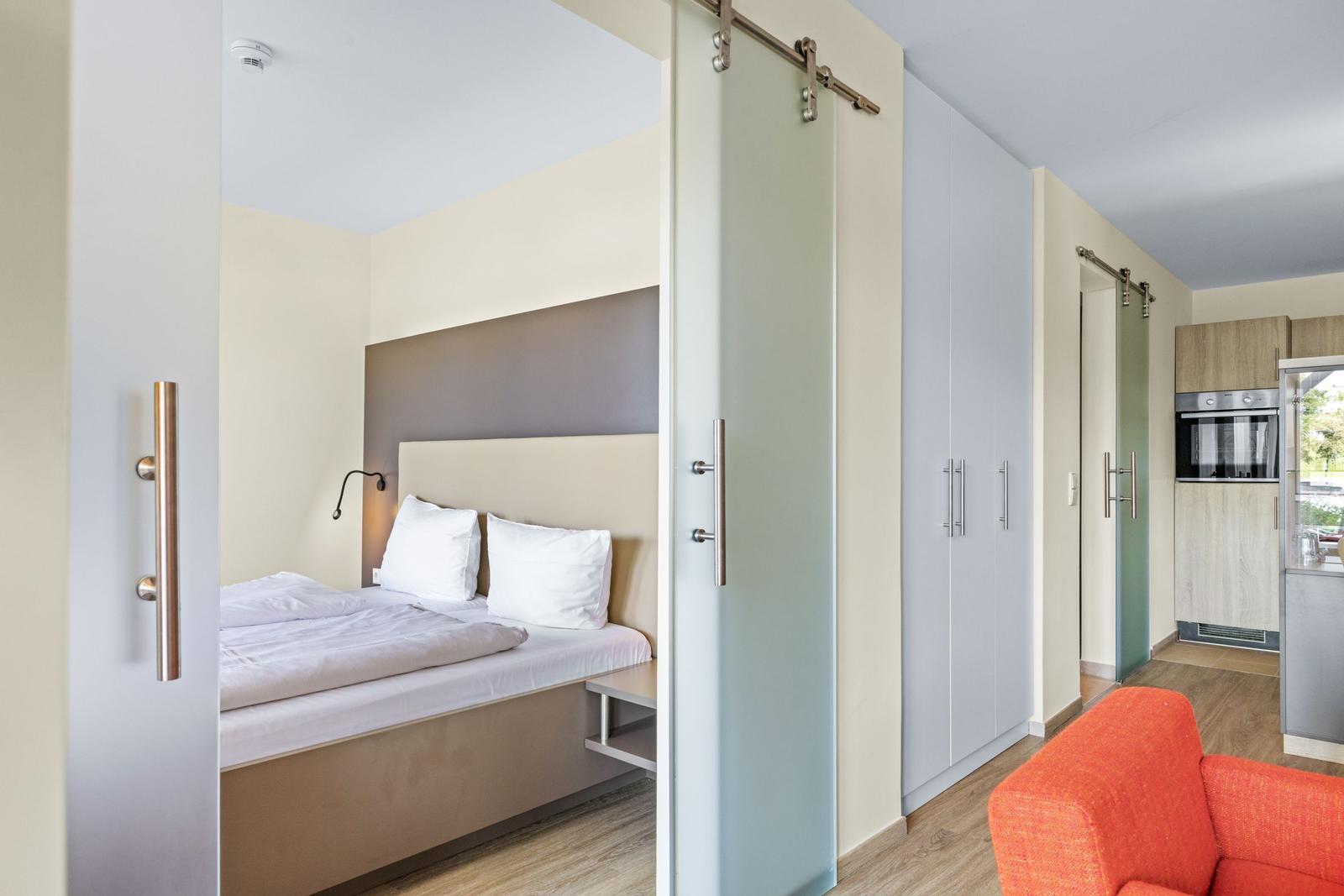 Schlafbereich mit Bett und Schiebetüren. Küche mit Kühlschrank und Ofen. Orange Sitzgelegenheit.