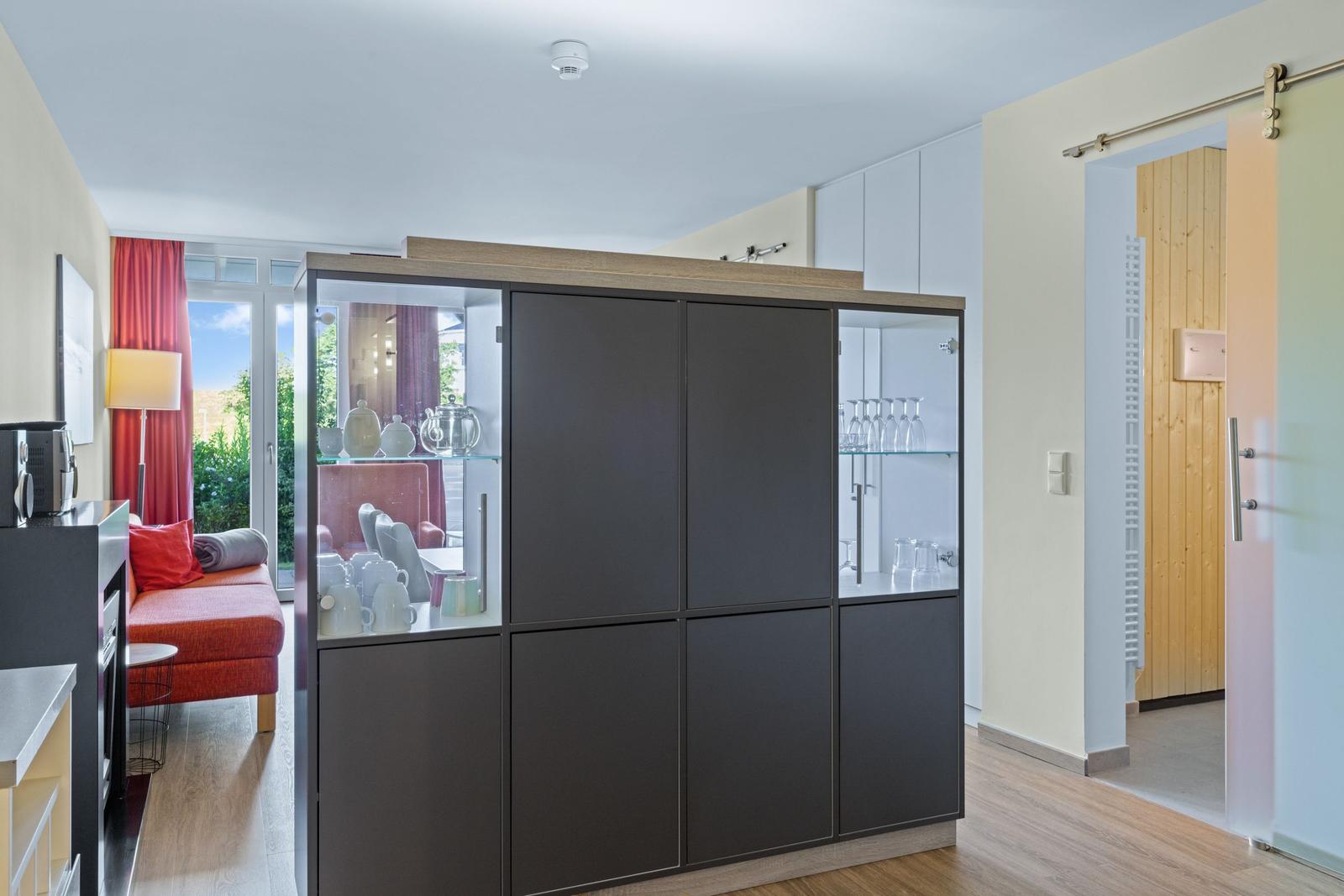 Großer Schrank mit Glasfront und Geschirr. Rote Couch und Holzboden. Tür zum Bad.