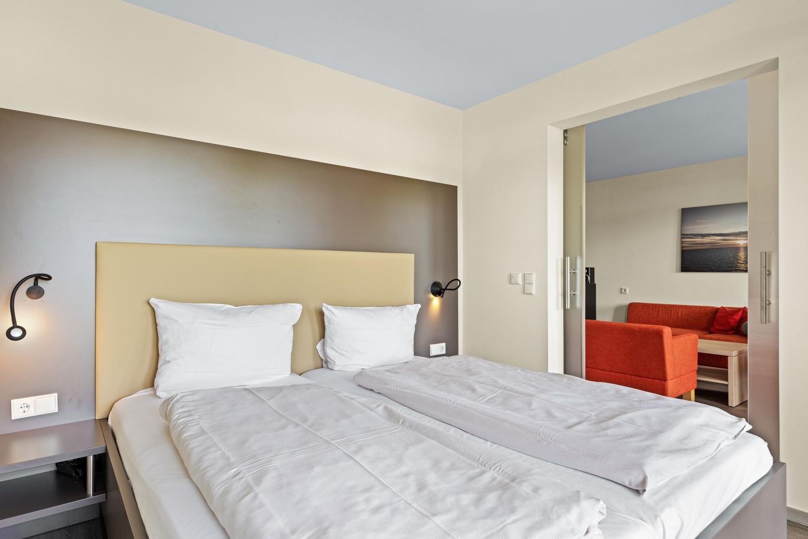 Schlafzimmer mit Doppelbett und Sicht auf Wohnbereich mit rotem Sofa.