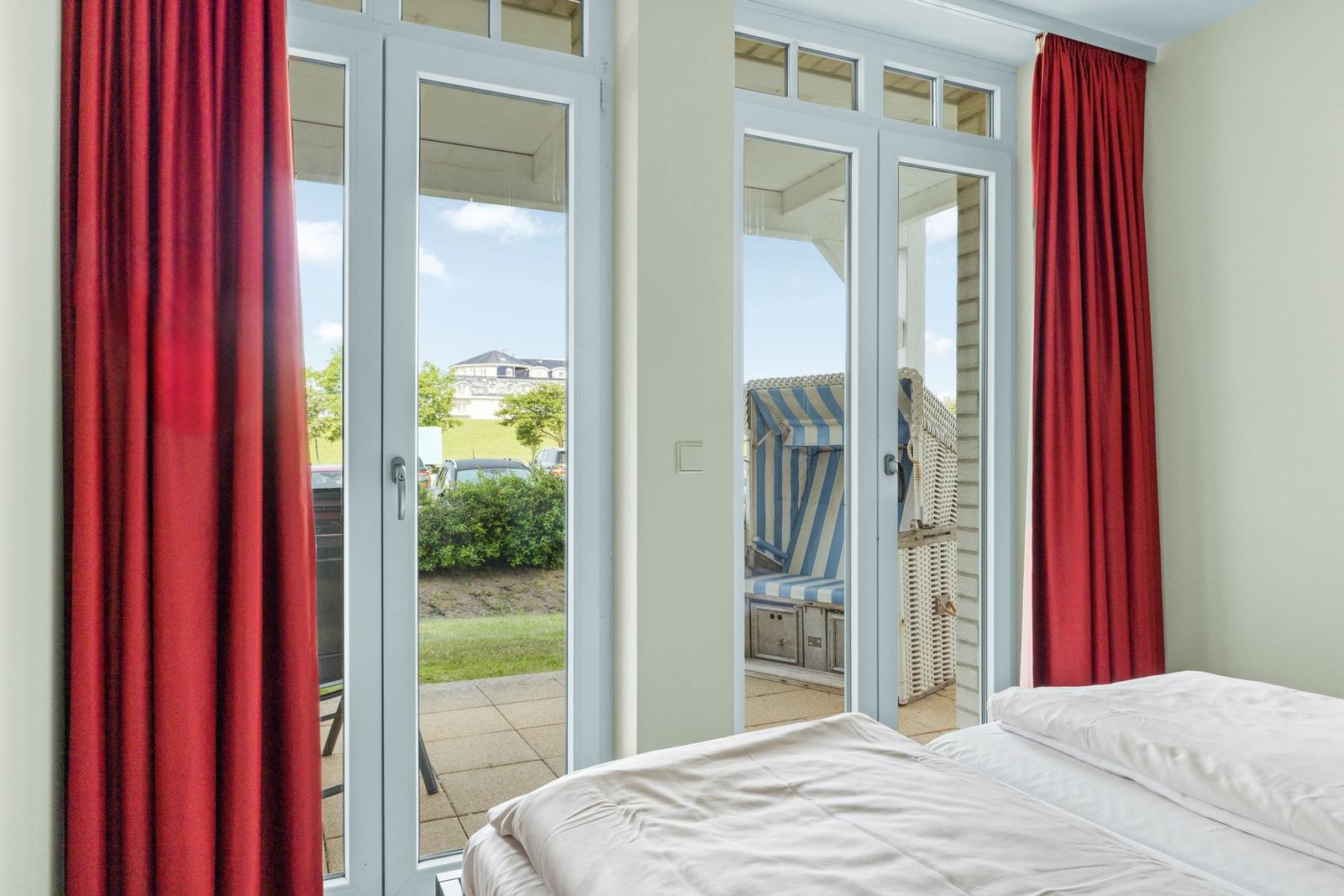 Zimmer mit Bett, roten Vorhängen und Blick auf Balkon mit Strandkorb.